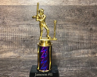 May include: Gold baseball trophy with a player figurine on top. The base is black with a purple and blue iridescent column. The base is engraved with "Jameson #11 Rockies 2022".