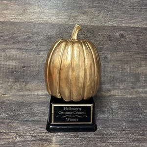 May include: A gold-colored pumpkin trophy with a black base. The trophy reads "Halloween Costume Contest Winner" in white text on a black plaque. The pumpkin has vertical ridges and a stem.