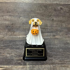 May include: A golden retriever dog dressed as a ghost holding a jack-o-lantern. The dog is on a black base with a gold plaque that reads "Pumpkin Carving Contest Winner".