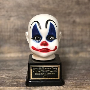 May include: A white porcelain clown head trophy with blue and red makeup, a black base, and a gold plaque that reads "Boyle Halloween Party Best Boy Costume 2022".