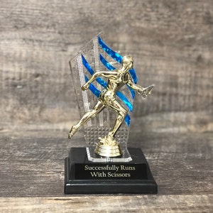 May include: A gold-colored trophy featuring a running figure holding scissors, set against a blue and clear backdrop. The trophy sits on a black base with the inscription "Successfully Runs With Scissors."