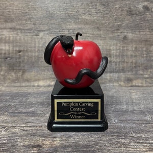 May include: A trophy featuring a red apple encircled by a black snake, mounted on a black base. The base has a gold plaque that reads "Pumpkin Carving Contest Winner."