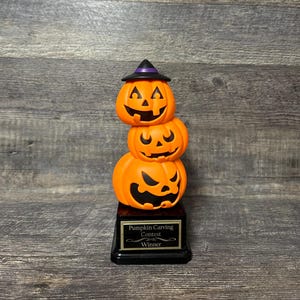 May include: A trophy featuring a stack of three orange jack-o'-lanterns with black carved faces, topped with a black and purple witch hat. The stack sits on a black rectangular base with a gold plaque that reads Pumpkin Carving Contest Winner.