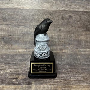 May include: A black crow figurine perched on a gray stone-textured pillar with skull carvings and the word SPOOKY embossed on the front. The pillar sits on a black square base with a gold-trimmed plaque reading Pumpkin Carving Contest Winner.