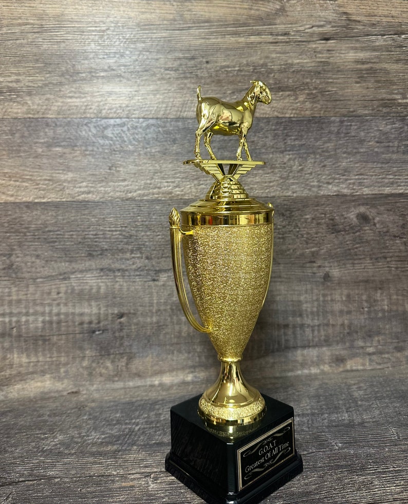 May include: A gold trophy with a goat figurine on top. The trophy has a black base with a plaque that reads "G.O.A.T. Greatest Of All Time".