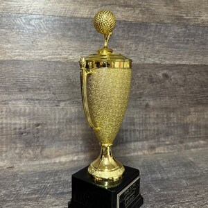 Może przedstawiać: Złoty puchar golfowy z czarną podstawą. Puchar ma na szczycie piłkę golfową i napis "Hole in One" z datą.