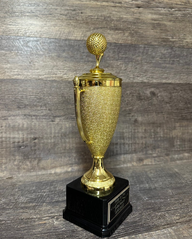 Może przedstawiać: Złoty puchar golfowy z czarną podstawą. Puchar ma na szczycie piłkę golfową i napis "Hole in One" z datą.