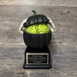 Puede incluir: Trofeo de concurso de disfraces de Halloween negro y verde con un cerebro dentro de una calabaza. La base del trofeo tiene una placa dorada que dice "Concurso de disfraces de Halloween Ganador".
