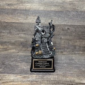 May include: A black and gold Halloween costume contest trophy featuring a spooky house with a tree, steps, and pumpkins. The base of the trophy reads "Halloween Costume Contest Winner".