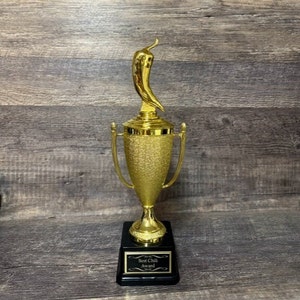 Può includere: Un trofeo dorato con un peperoncino in cima. La base del trofeo è nera con una placca dorata che recita "Best Chili Award".