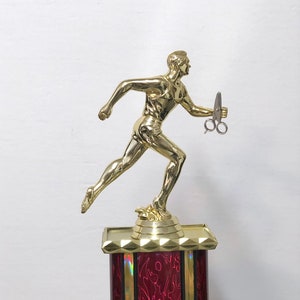 May include: Gold-colored trophy with a running figure holding a pair of scissors. The base of the trophy is red with a black and gold trim.