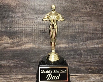May include: A gold-colored trophy shaped like a person holding a heart. The trophy is on a black base with a plaque that reads "World's Greatest Dad".