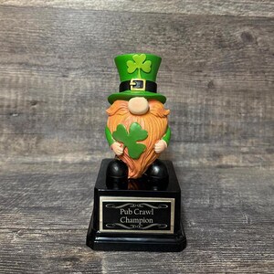 Puede incluir: Una figurita de duende verde sosteniendo un trébol, con un sombrero verde con una hebilla dorada, de pie sobre una base negra con una placa dorada que dice "Pub Crawl Champion".