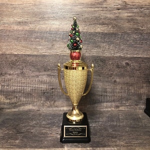 May include: A gold-colored trophy with a textured body and two handles. The trophy is topped with a small Christmas tree decorated with red, green, and gold ornaments. The base is black with a plaque that reads "Most Holiday Spirit."