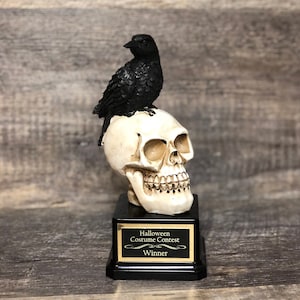 May include: A black crow perched on a white skull trophy with a black base. The base has a gold plaque that reads "Halloween Costume Contest Winner".