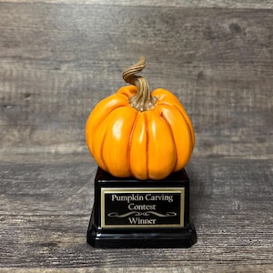 Puede incluir: Un trofeo para el ganador de un concurso de tallado de calabazas. La calabaza naranja se asienta sobre una base negra con una placa dorada que dice "Pumpkin Carving Contest Winner". La calabaza tiene un tallo marrón.