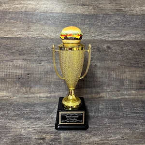 May include: A gold-colored trophy with a black base, topped with a realistic burger. The trophy has a glittery finish and curved handles. The base has a plaque that reads "You Deserve A Trophy".