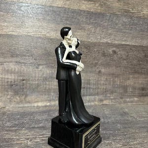 May include: Black and white skeletal couple figurine in a wedding pose. The couple is standing on a black base with a gold inscription that reads "Rest In Peace Together".