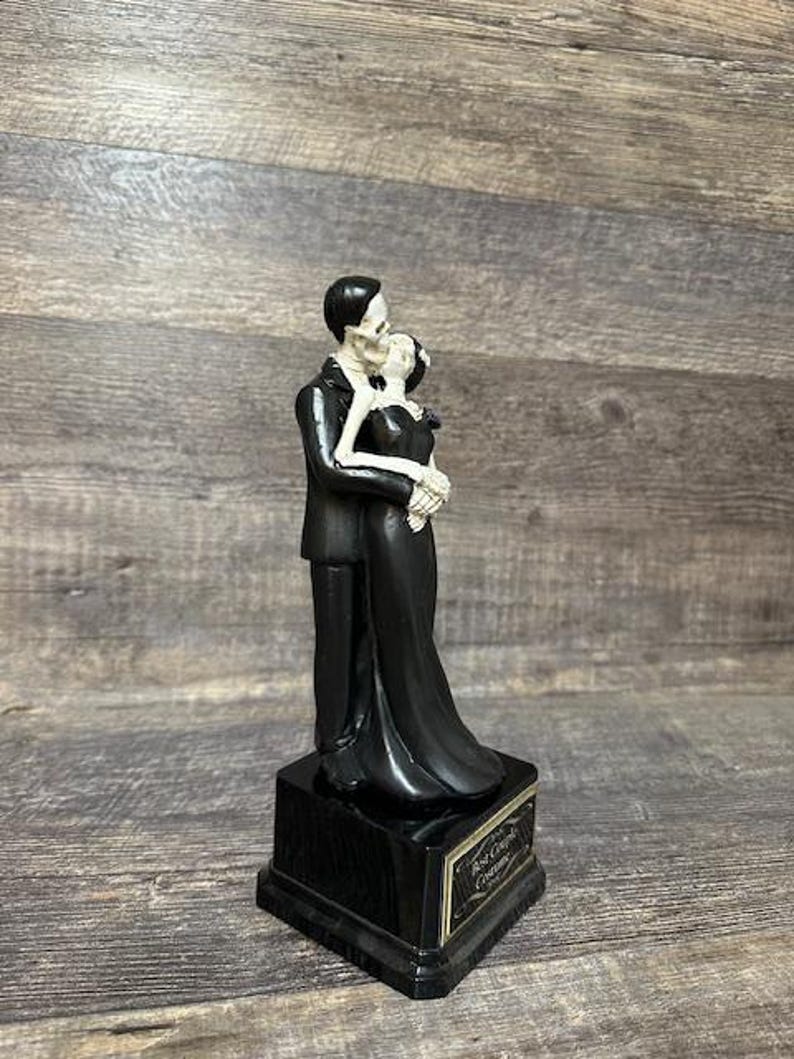 May include: Black and white skeletal couple figurine in a wedding pose. The couple is standing on a black base with a gold inscription that reads "Rest In Peace Together".