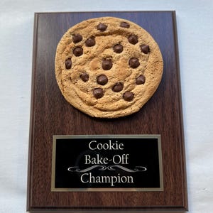 May include: A chocolate chip cookie trophy on a dark wood plaque. The cookie is round, with visible chocolate chips. A black plate below reads "Cookie Bake-Off Champion" in white text.