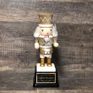 May include: A gold and white glitter nutcracker holding drumsticks and a drum. The nutcracker is standing on a black base with a gold plaque that reads "Ugliest Sweater".