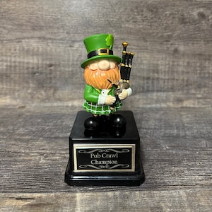 Puede incluir: Una figurita de duende verde tocando una gaita sobre una base negra con una placa dorada que dice "Pub Crawl Champion".