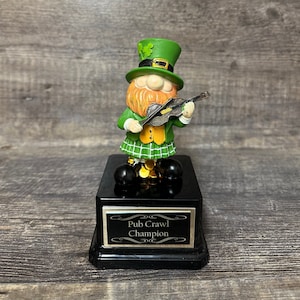 Puede incluir: Un trofeo negro con una placa dorada que dice "Pub Crawl Champion". Una figura de duende verde con barba roja y un violín está en la parte superior del trofeo.