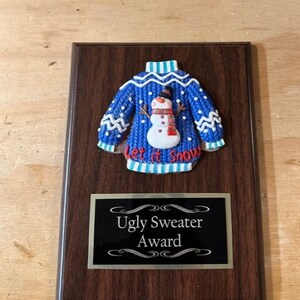 May include: A wooden plaque with a blue and white 3D ugly Christmas sweater with a snowman and the text "Let it Snow". The plaque has a gold border and a black background with the text "Ugly Sweater Award".