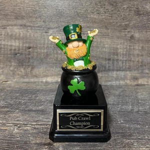 Puede incluir: Un trofeo del Día de San Patricio con un duende que emerge de una olla de oro. El duende lleva un sombrero y una chaqueta verdes. La base negra del trofeo dice "Pub Crawl Champion".