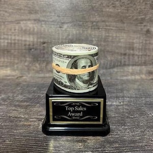 May include: A black award plaque with a gold trim and the text "Top Sales Award". A rolled up stack of fake money with a rubber band around it sits on top of the plaque.