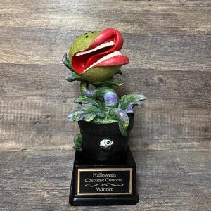 Puede incluir: Un trofeo de ganador del concurso de disfraces de Halloween. Presenta una planta verde con una gran boca abierta y labios rojos, colocada en una maceta negra con un ojo. El trofeo está sobre una base negra con una placa dorada que dice "Halloween Costume Contest Winner."