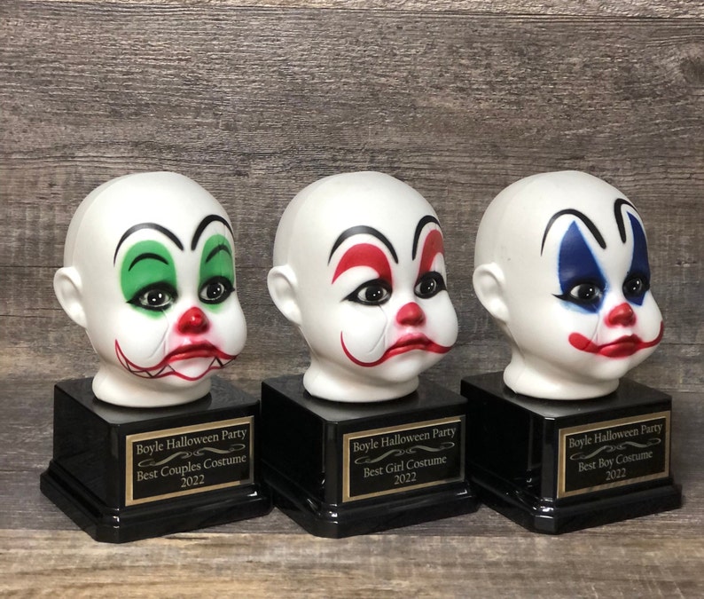 May include: Three black and gold award bases with white clown head figurines. The bases have gold plaques with the text "Boyle Halloween Party" and the categories "Best Couples Costume", "Best Girl Costume", and "Best Boy Costume".