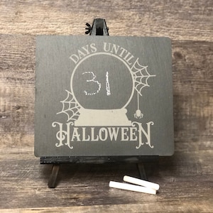 May include: A black chalkboard with white text that reads "Days Until Halloween". A white circle with a spider web design and the number 31 written in white chalk is in the center of the chalkboard. The chalkboard is on a small wooden easel.