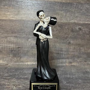 May include: A black and white skeleton couple figurine on a black base with a gold inscription that reads "Best Couple Costume".
