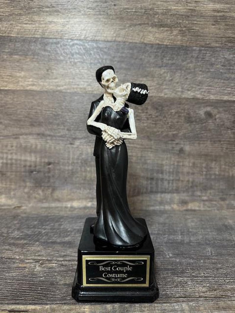 May include: A black and white skeleton couple figurine on a black base with a gold inscription that reads "Best Couple Costume".