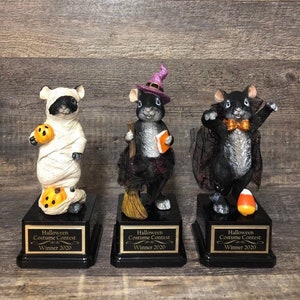 May include: Three black and white mouse figurines dressed in Halloween costumes. The mouse on the left is dressed as a mummy, the middle mouse is dressed as a witch, and the mouse on the right is dressed as a vampire. Each mouse is standing on a black base with a gold plaque that reads "Halloween Costume Contest Winner 2020".