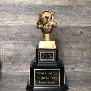 Puede incluir: Un trofeo negro y dorado con una parte superior en forma de inodoro. La base del trofeo tiene una placa negra con el texto "Fantasy Football" y una segunda placa con el texto "Your Custom Logo & Text Goes Here".
