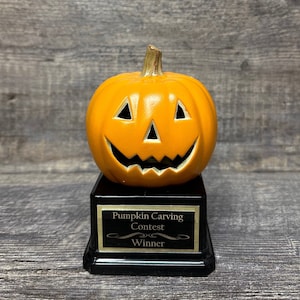 May include: A black and gold pumpkin carving contest trophy with a smiling orange pumpkin on top. The plaque reads "Pumpkin Carving Contest Winner".