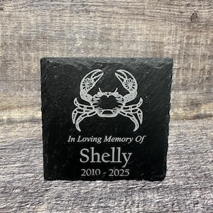 Puede incluir: Placa conmemorativa de pizarra negra con un diseño de cangrejo blanco, el texto "In Loving Memory Of Shelly 2010 - 2025" está grabado en la placa.