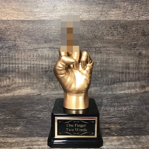 May include: A gold-colored trophy with a hand giving the middle finger. The base of the trophy is black with a gold plaque that reads "One Finger Two Words".