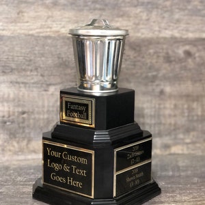 May include: A black trophy with a silver trash can on top. The trophy has three tiers with gold lettering. The top tier says "Fantasy Football". The middle tier says "Your Custom Logo & Text Goes Here". The bottom tier has the text "2019 Zach Grimes (2-11)" and "2018 Shawn Smith (3-10)".