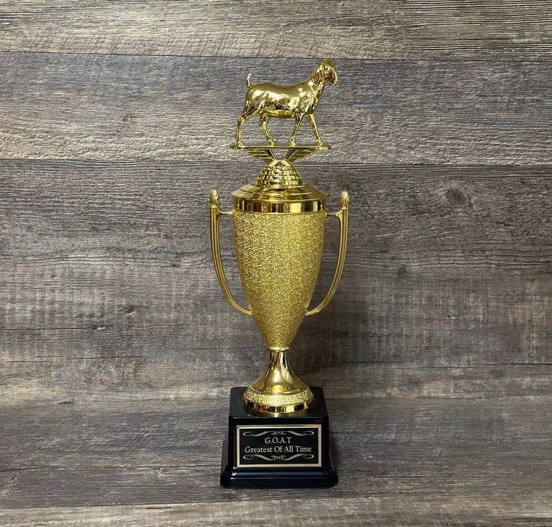 May include: A gold trophy with a goat figurine on top. The trophy has a black base with a gold plaque that reads "GOAT Greatest Of All Time".