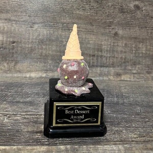 May include: A black award plaque with a gold border and the text "Best Dessert Award". A chocolate ice cream cone trophy sits on top of the plaque.