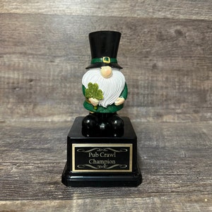 Könnte beinhalten: Eine schwarze und grüne Gnome-Trophäe mit einem goldenen Gürtel und einem Kleeblatt in der Hand. Der Sockel der Trophäe ist schwarz mit einer goldenen Plakette, auf der "Pub Crawl Champion" steht.