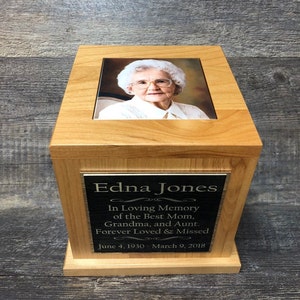 May include: A wooden cremation urn with a photo of a woman on the top and a brass plaque with the text "Edna Jones In Loving Memory of the Best Mom, Grandma, and Aunt. Forever Loved & Missed June 4, 1930 - March 9, 2018"