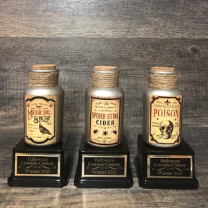 Puede incluir: Tres botellas de trofeo temáticas de Halloween con tapones de corcho y detalles de cuerda. Cada botella tiene una etiqueta de estilo vintage con textos como "Medicine Show", "Spider Sting Cider" y "Poison". Las botellas están sobre bases negras con placas "Halloween Costume Contest Winner 2020".