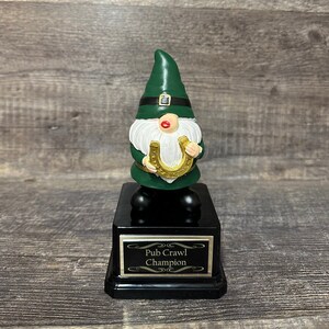 Könnte beinhalten: Ein grüner Zwerg, der ein goldenes Hufeisen hält, mit einer schwarzen Basis und einer goldenen Plakette mit der Aufschrift "Pub Crawl Champion".