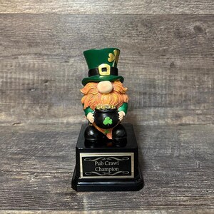 Puede incluir: Figura de duende con sombrero de copa verde y barba naranja, sosteniendo una olla de oro con un trébol. La figura está sobre una base negra con el texto "Pub Crawl Champion".