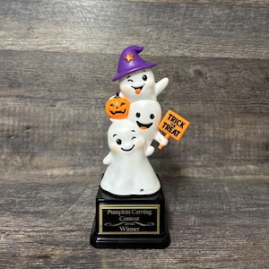 May include: A trophy featuring three stacked white ghost figurines. The top ghost wears a purple witch hat with a gold star. The middle ghost holds an orange jack-o'-lantern and a sign that reads TRICK OR TREAT. The black base has a plaque reading Pumpkin Carving Contest Winner.