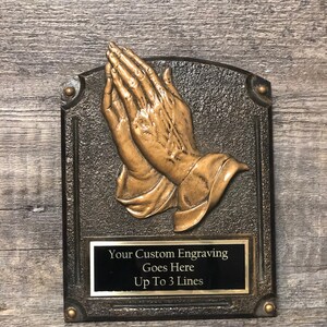 May include: A bronze plaque with a raised image of two hands clasped in prayer. The plaque has a black rectangular plate with the text "Your Custom Engraving Goes Here Up To 3 Lines".